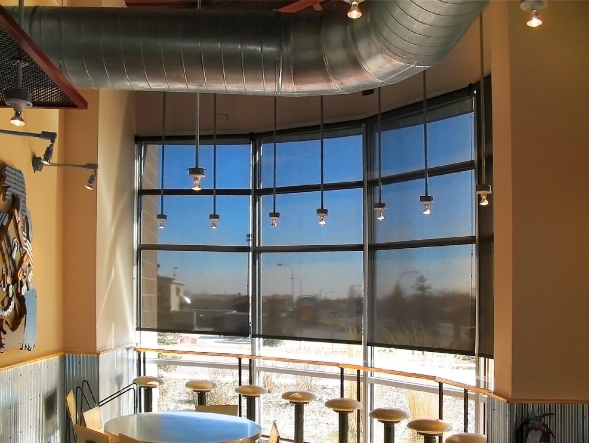 Commercial solar shades on floor-to-ceiling windows in a restaurant with exposed ductwork